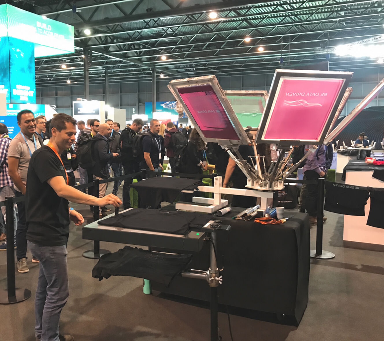 An attendee screen prints a black t-shirt with a pink Be Data Driven design as a queue forms at a live screen printing stand in a Barcelona conference hall